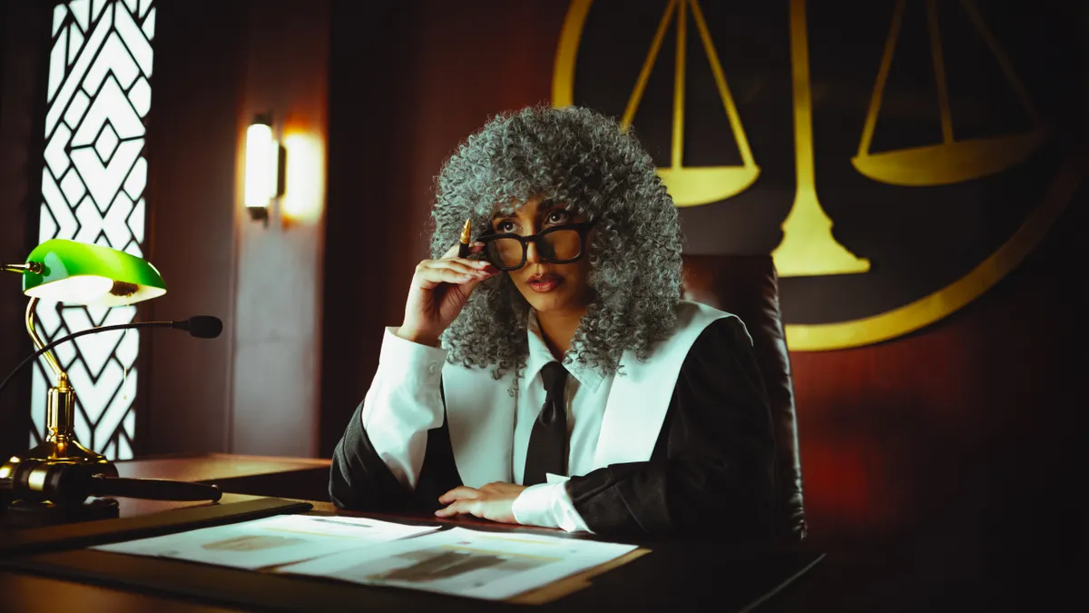 Judge in traditional attire sits at a courtroom desk with a serious expression