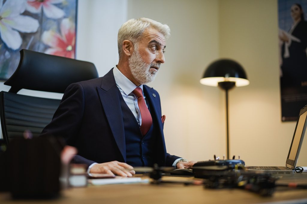 A gray-haired man in a suit sits at a desk in a modern office